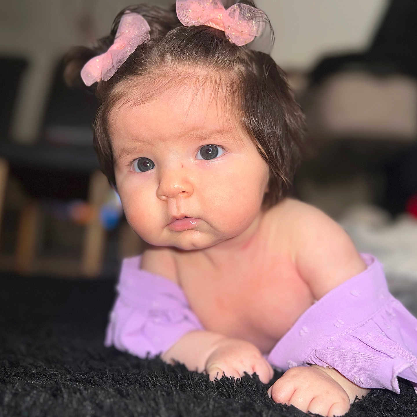 Emmy is registered to the contest to win money with this photo: baby, bows, carpet, child, curious, cute, expression, eyes, face, floor, hair, indoor, infant, lying_down, person, portrait, purple_clothing, skin, soft_texture, young_child