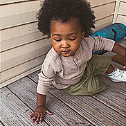 Luca is registered to the contest to win money with this photo: child, toddler, afro_hair, curly_hair, long_sleeve_shirt, shorts, sneakers, wooden_floor, porch, outdoor, skin, person, young_child, candid, casual_clothing, brown_skin, exploring, daylight, natural_light, contemplative
