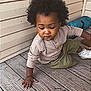 child, toddler, afro_hair, curly_hair, long_sleeve_shirt, shorts, sneakers, wooden_floor, porch, outdoor, skin, person, young_child, candid, casual_clothing, brown_skin, exploring, daylight, natural_light, contemplative