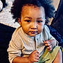 toddler, child, afro_hair, curly_hair, beige_shirt, green_shorts, gold_chain, sitting, indoor, portrait, cute, young_child, looking_at_camera, skin, hand, face, expression, casual_clothing, bed, blanket