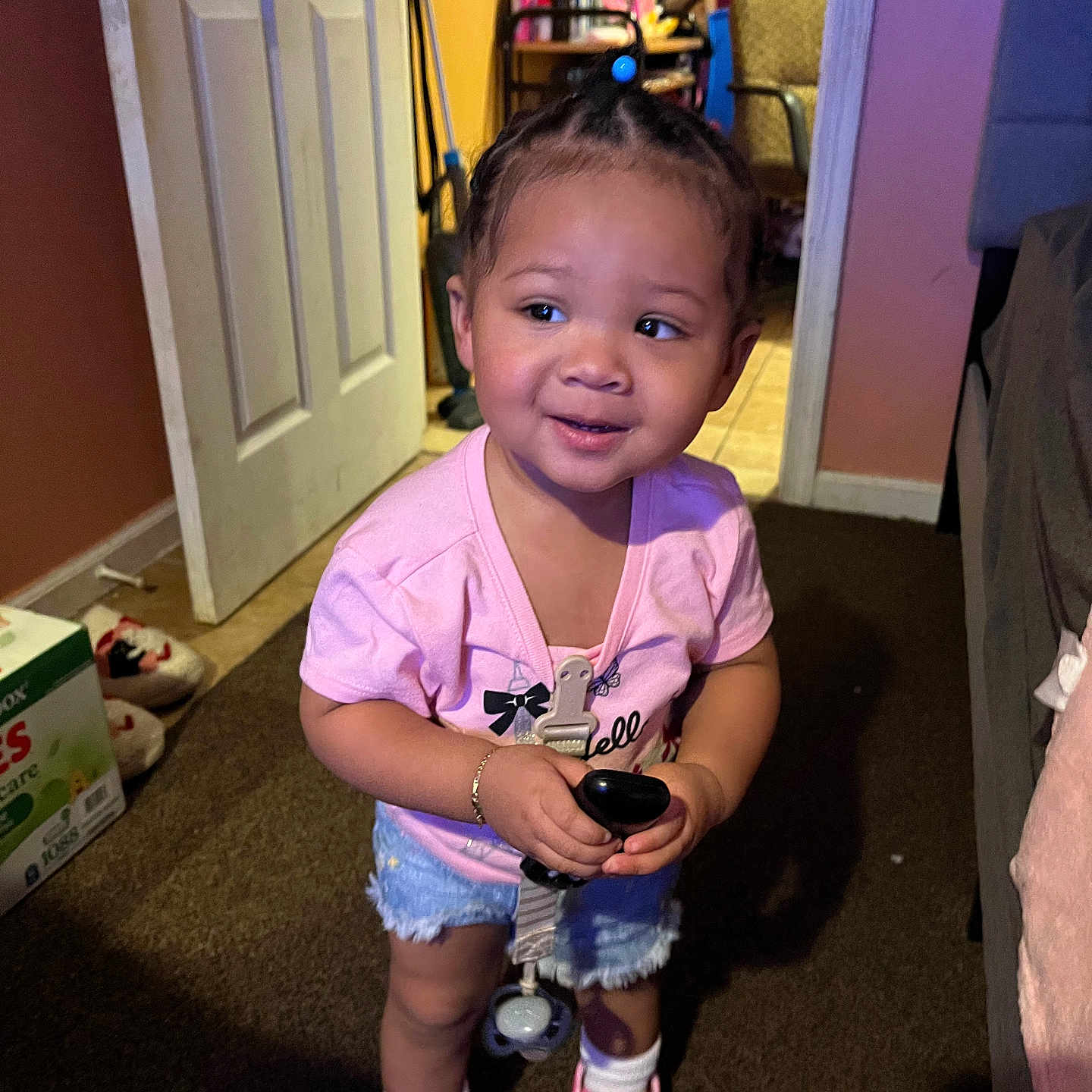 Hadassah joined the competition — help win amazing prizes! toddler, child, indoor, carpet, pink_shirt, denim_shorts, white_socks, pink_shoes, holding_object, curious_expression, door, chair, household_items, floor, person, shorts, footwear, hair_tied, standing, casual_clothing
