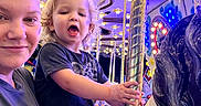 Kolton is registered to the contest to win money with this photo: child, toddler, adult, carousel, horse, amusement_ride, carnival_lights, smile, blonde_hair, curly_hair, blue_shorts, black_shirt, golden_pole, fun, outdoor, night, family, joy, portrait, face