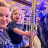 child, toddler, adult, carousel, horse, amusement_ride, carnival_lights, smile, blonde_hair, curly_hair, blue_shorts, black_shirt, golden_pole, fun, outdoor, night, family, joy, portrait, face