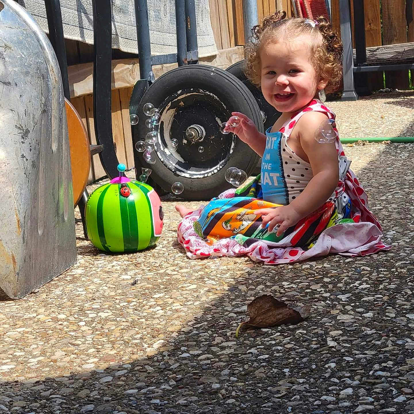 Scarlet joined the competition — help win amazing prizes! bubbles, child, colorful, curly_hair, daytime, dress, fence, garden, ground, happy, outdoor, patio, person, play, smile, summer, sunlight, toddler, toy, wheelbarrow