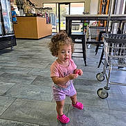 Scarlet is registered to the contest to win money with this photo: toddler, child, curly_hair, pink_clothing, pink_shoes, indoor, tile_floor, shopping_cart, clothing_rack, counter, store_interior, person, holding_object, casual_wear, natural_light, door, window, flooring, furniture, shopping