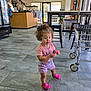 toddler, child, curly_hair, pink_clothing, pink_shoes, indoor, tile_floor, shopping_cart, clothing_rack, counter, store_interior, person, holding_object, casual_wear, natural_light, door, window, flooring, furniture, shopping