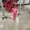 child, girl, pink_clothing, boots, wheel, red_wheel, museum_display, glass_case, floor, reflection, stanchion, indoor, braided_hair, curious, standing, exhibit, tile_floor, casual_wear, young_person, display_case