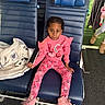 child, pink_clothing, boots, blue_chair, stuffed_animal, indoor, carpet, people, seated, serious_expression, braided_hair, patterned_pants, casual_wear, flooring, holding_armrest, waiting_area, group, multicolor, young_child, portrait