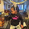 child, birthday, headband, black_hoodie, puma, table, living_room, decorations, banner, window, sofa, pottery, patterned_pants, celebration, indoor, person, furniture, curtains, light, home