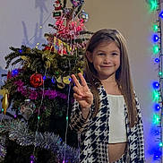Ilanie participe au concours pour gagner de l'argent avec cette photo : celebration, child, christmas_tree, decorations, festive, girl, happy, holiday_lights, indoor, long_hair, ornaments, patterned_clothing, peace_sign, person, photography, shadow, smile, star, tinsel, wall