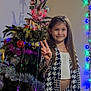 celebration, child, christmas_tree, decorations, festive, girl, happy, holiday_lights, indoor, long_hair, ornaments, patterned_clothing, peace_sign, person, photography, shadow, smile, star, tinsel, wall