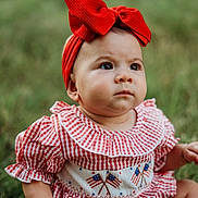 Alexandra joined the competition — help win amazing prizes! baby, child, red_bow, headband, checkered_clothing, grass, outdoor, sitting, portrait, cute, infant, young_child, summer, nature, face, expression, hands, legs, patterned_clothing, background_blur
