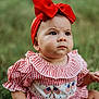 baby, child, red_bow, headband, checkered_clothing, grass, outdoor, sitting, portrait, cute, infant, young_child, summer, nature, face, expression, hands, legs, patterned_clothing, background_blur