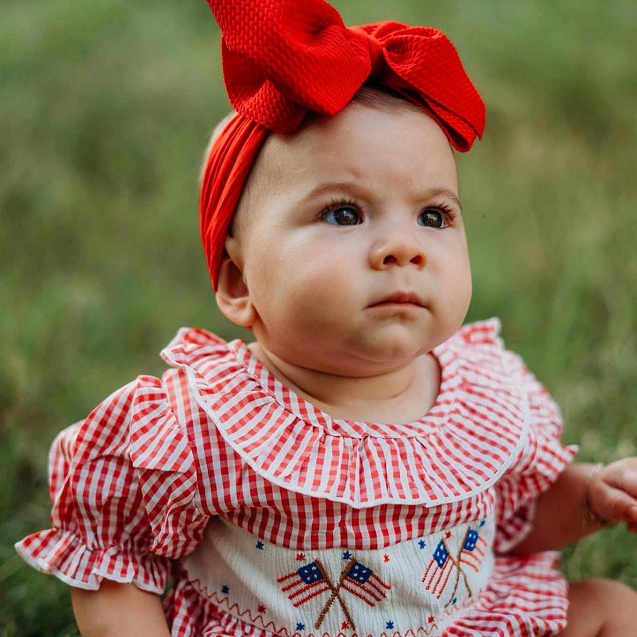 Alexandra joined the competition — help win amazing prizes! baby, background_blur, checkered_clothing, child, cute, expression, face, grass, hands, headband, infant, legs, nature, outdoor, patterned_clothing, portrait, red_bow, sitting, summer, young_child