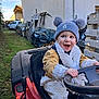 toddler, child, hat, pom_poms, scarf, ride_on_toy, steering_wheel, red_vehicle, outdoor, grass, boots, jacket, happy, smiling, yard, tools, car, fence, wood, playtime