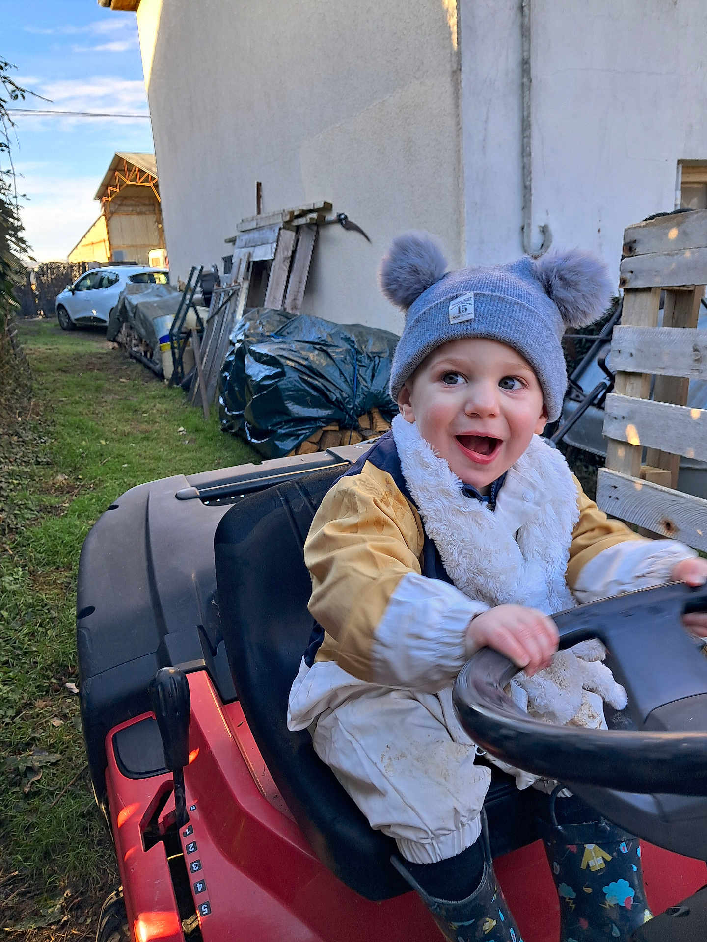 Ayden participe au concours pour gagner de l'argent avec cette photo : toddler, child, hat, pom_poms, scarf, ride_on_toy, steering_wheel, red_vehicle, outdoor, grass, boots, jacket, happy, smiling, yard, tools, car, fence, wood, playtime
