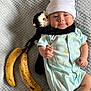baby, infant, plush_toy, monkey_toy, banana, onesie, hat, blanket, smiling, cute, lying_down, soft_toy, baby_clothing, cozy, indoor, child, happy, toy, fruit, portrait