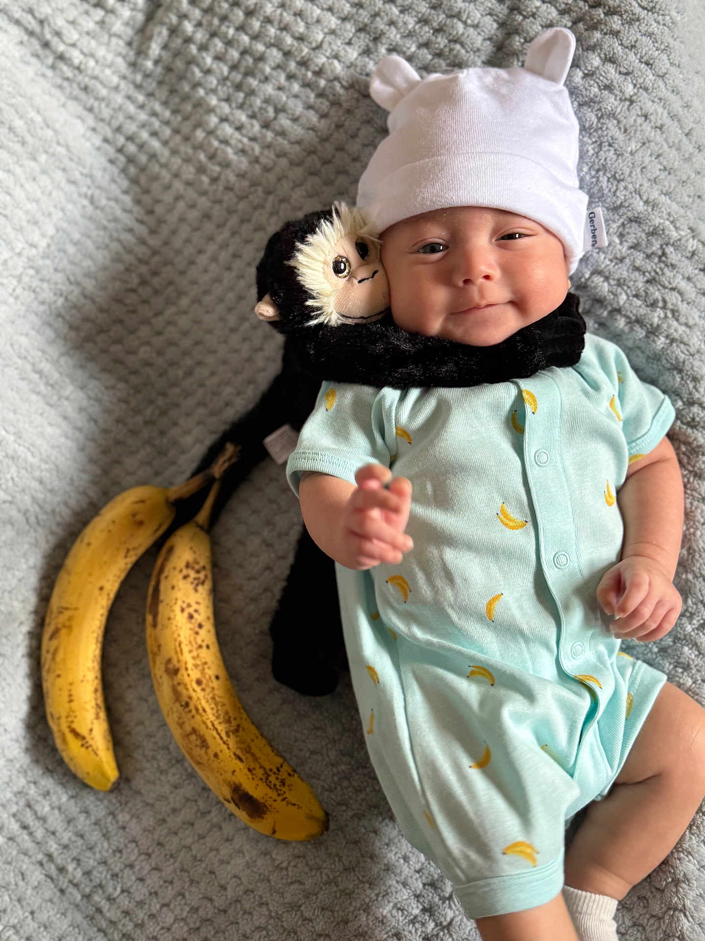Kavion joined the competition — help win amazing prizes! baby, infant, plush_toy, monkey_toy, banana, onesie, hat, blanket, smiling, cute, lying_down, soft_toy, baby_clothing, cozy, indoor, child, happy, toy, fruit, portrait