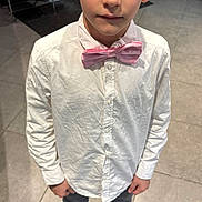 Tiago participe au concours pour gagner de l'argent avec cette photo : child, boy, white_shirt, bow_tie, pink_bow_tie, indoor, tile_floor, barefoot, socks, standing, looking_up, blue_eyes, short_hair, clothing, casual, portrait, person, furniture, chair, background