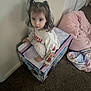 blanket, box, carpet, child, clothing, curious, cute, door, floor, frozen_theme, home, indoor, looking_up, pet_bed, pigtails, soft_lighting, toddler, toy_box, wall, young_child