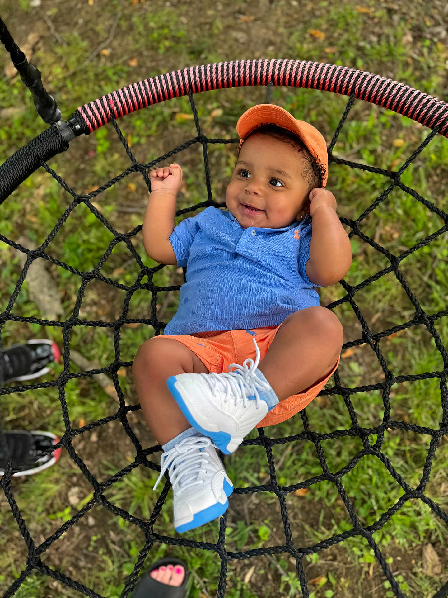 Traesyn is registered to the contest to win money with this photo: baby, blue_shirt, casual, child, cute, feet, footwear, grass, happy, leisure, net_swing, orange_cap, orange_shorts, outdoor, person, play, smiling, summer, swing, white_sneakers