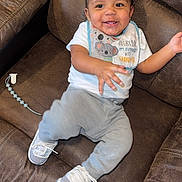 Traesyn is registered to the contest to win money with this photo: baby, bib, brown_furniture, child, couch, cozy, curly_hair, cute, face, footwear, gray_pants, hand, happy, indoor, person, playful, seated, smiling, sneakers, white_shirt