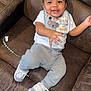 baby, bib, brown_furniture, child, couch, cozy, curly_hair, cute, face, footwear, gray_pants, hand, happy, indoor, person, playful, seated, smiling, sneakers, white_shirt