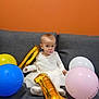 baby, child, balloon, gold_balloon, number_one, pacifier, couch, gray_couch, white_dress, sitting, celebration, birthday, indoor, orange_wall, pink_balloon, blue_balloon, yellow_balloon, cute, party, infant