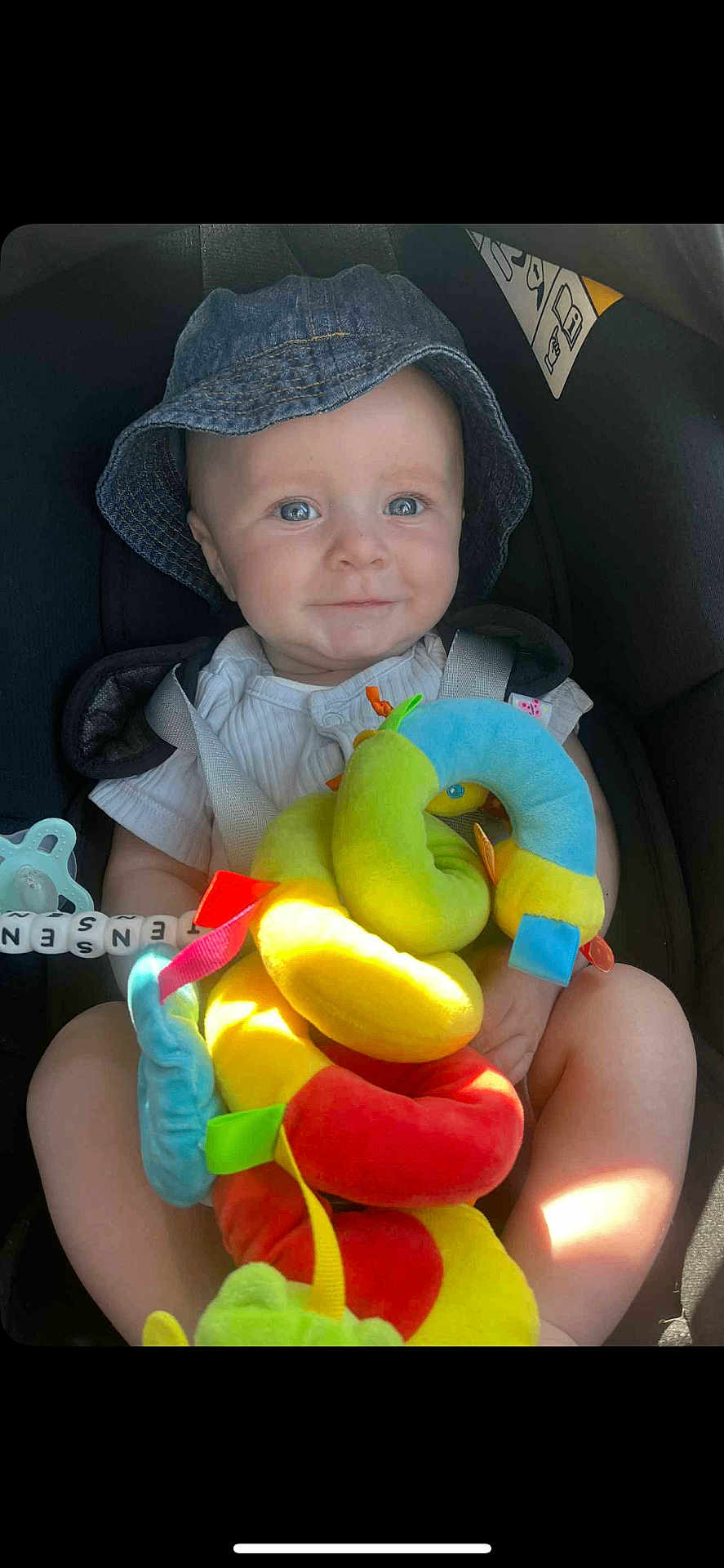 Jensen is registered to the contest to win money with this photo: baby, child, denim_hat, bucket_hat, car_seat, plush_toy, colorful_toy, soft_toy, straps, blue_eyes, cute, infant, smiling, indoor, seatbelt, child_safety, toy_snake, happy, person, closeup