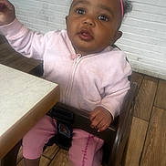 Malasianay is registered to the contest to win money with this photo: baby, child, infant, high_chair, pink_outfit, headband, bow, curly_hair, smiling, portrait, sitting, indoors, kitchen, table, wood_floor, brick_wall, sock, reaching, family_photo, cute