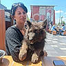 woman, puppy, dog, outdoor, cafe, table, sunny, daytime, people, black_sweater, fur, pet, holding, bench, building, sky, glasses, phone, collar, happy