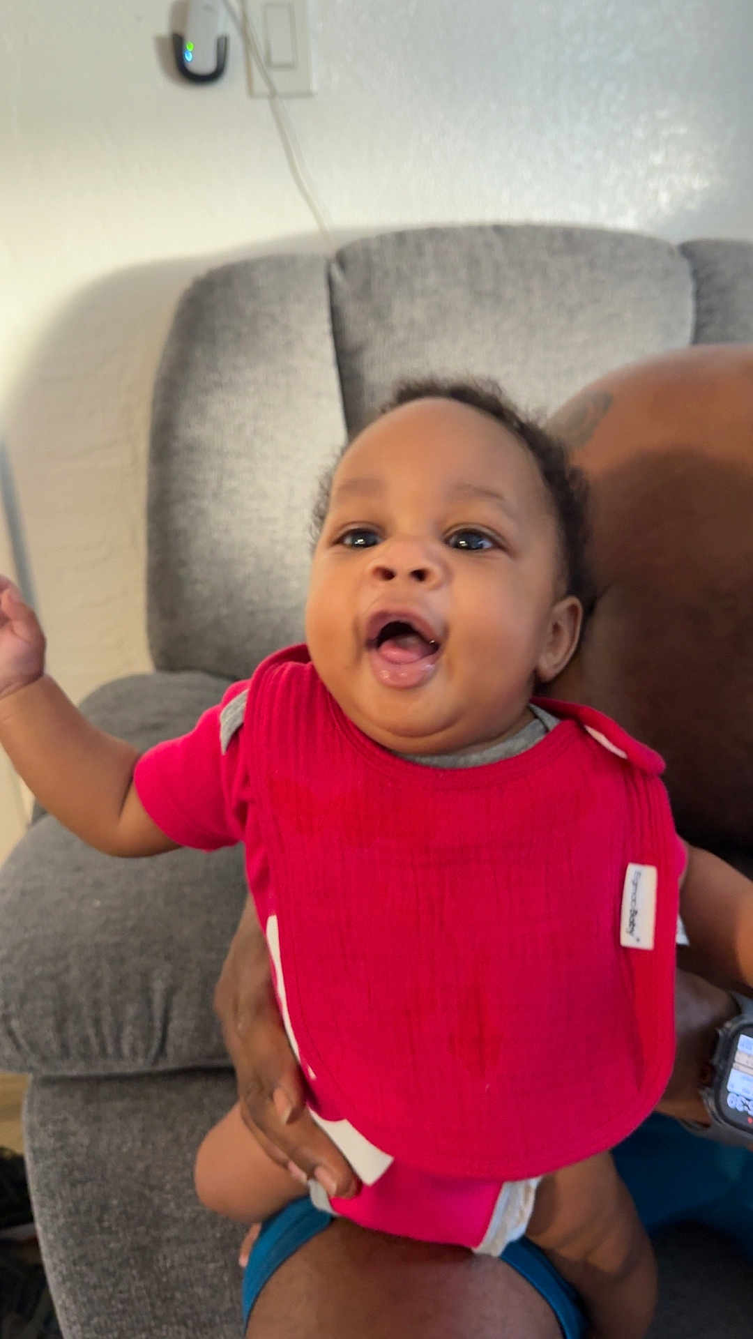 Zyaire is registered to the contest to win money with this photo: baby, infant, red_bib, smiling, person, indoor, couch, adult_hand, happy, child, home, seated, cute, skin, clothing, face, portrait, furniture, watch, expression