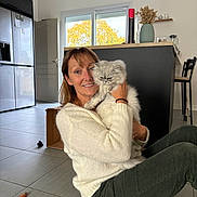 Athos participe au concours pour gagner de l'argent avec cette photo : woman, cat, indoor, kitchen, floor, sitting, smiling, fluffy_cat, cardboard_box, window, tree, sweater, corduroy_pants, cat_brush, tile_floor, modern_interior, vase, chair, table, door