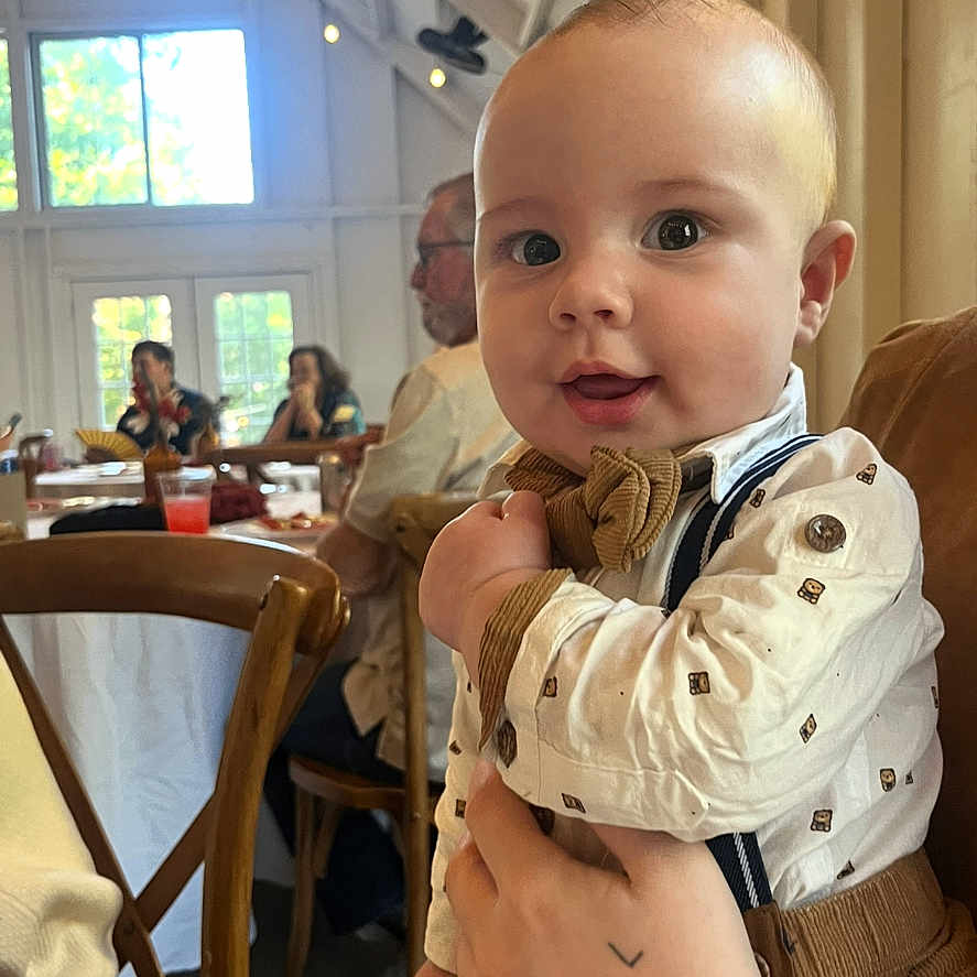 Julien is registered to the contest to win money with this photo: adult, baby, background, bow_tie, chair, child, clothing, curious, cute, face, furniture, hand, indoor, light, person, portrait, smile, suspenders, window, wood