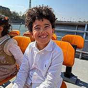 Milan participe au concours pour gagner de l'argent avec cette photo : child, boy, smiling, curly_hair, white_shirt, orange_seat, outdoor, daylight, eiffel_tower, river, happy, portrait, friends, bench, blue_sky, casual_clothing, tourist_spot, playful, sunny, urban