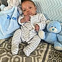baby, infant, striped_onesie, plush_elephant, blue_teddy_bear, blanket, pillow, patterned_bedspread, indoor, child, cute, soft_toys, seated, portrait, newborn, cozy, comfort, resting, small_hands, striped_clothing