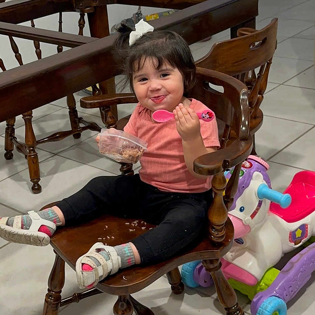 Zoé Isabella Villeda is registered to the contest to win money with this photo: bow, chair, child, clothing, food_container, furniture, hair_accessory, indoor, person, pink_spoon, playful, ride_on_toy, sandals, sitting, smiling, socks, tile_floor, toddler, toy_horse, wooden_chair