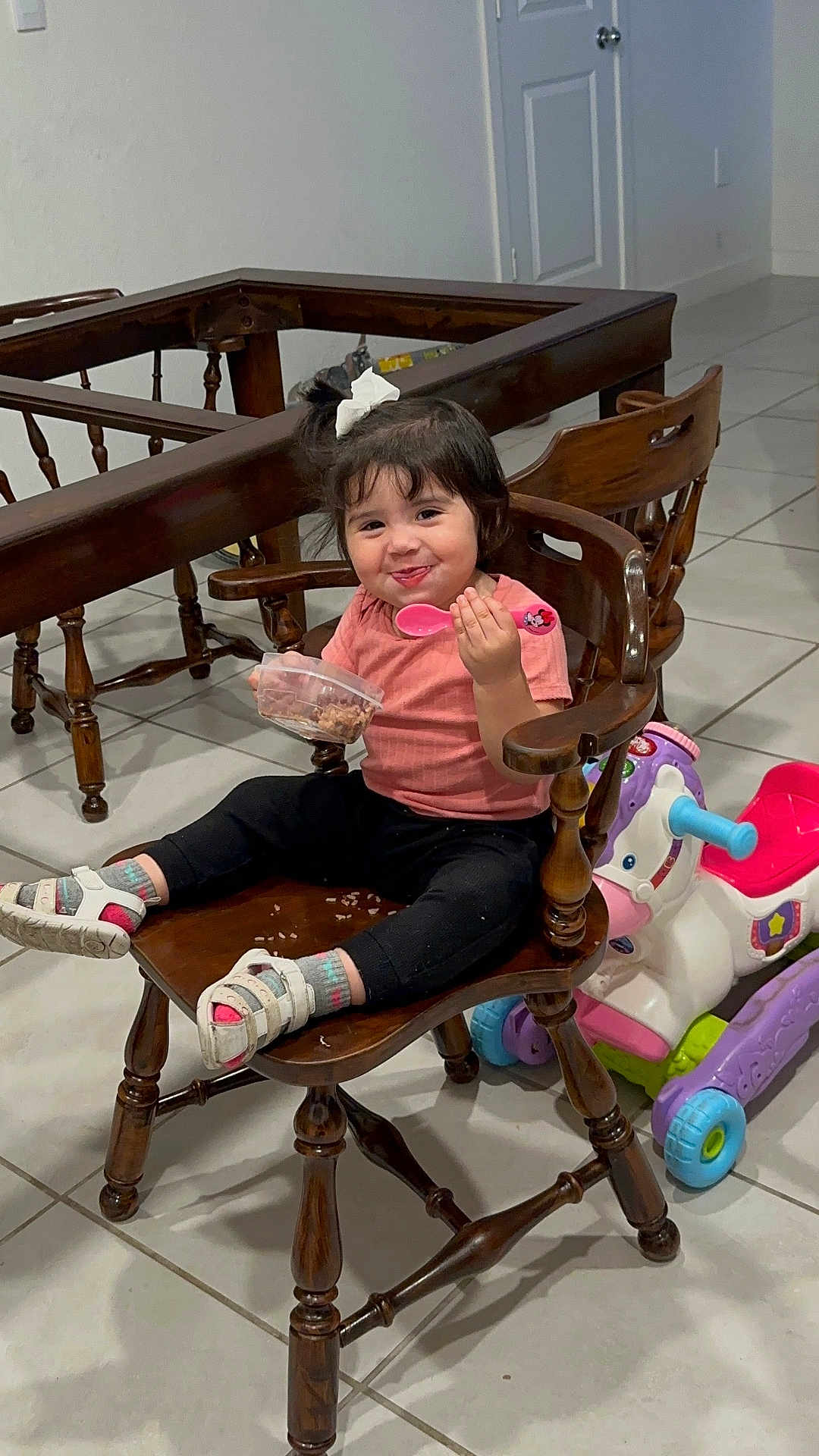Zoé Isabella Villeda is registered to the contest to win money with this photo: toddler, child, chair, wooden_chair, pink_spoon, food_container, bow, hair_accessory, ride_on_toy, toy_horse, indoor, tile_floor, smiling, sitting, clothing, socks, sandals, furniture, person, playful