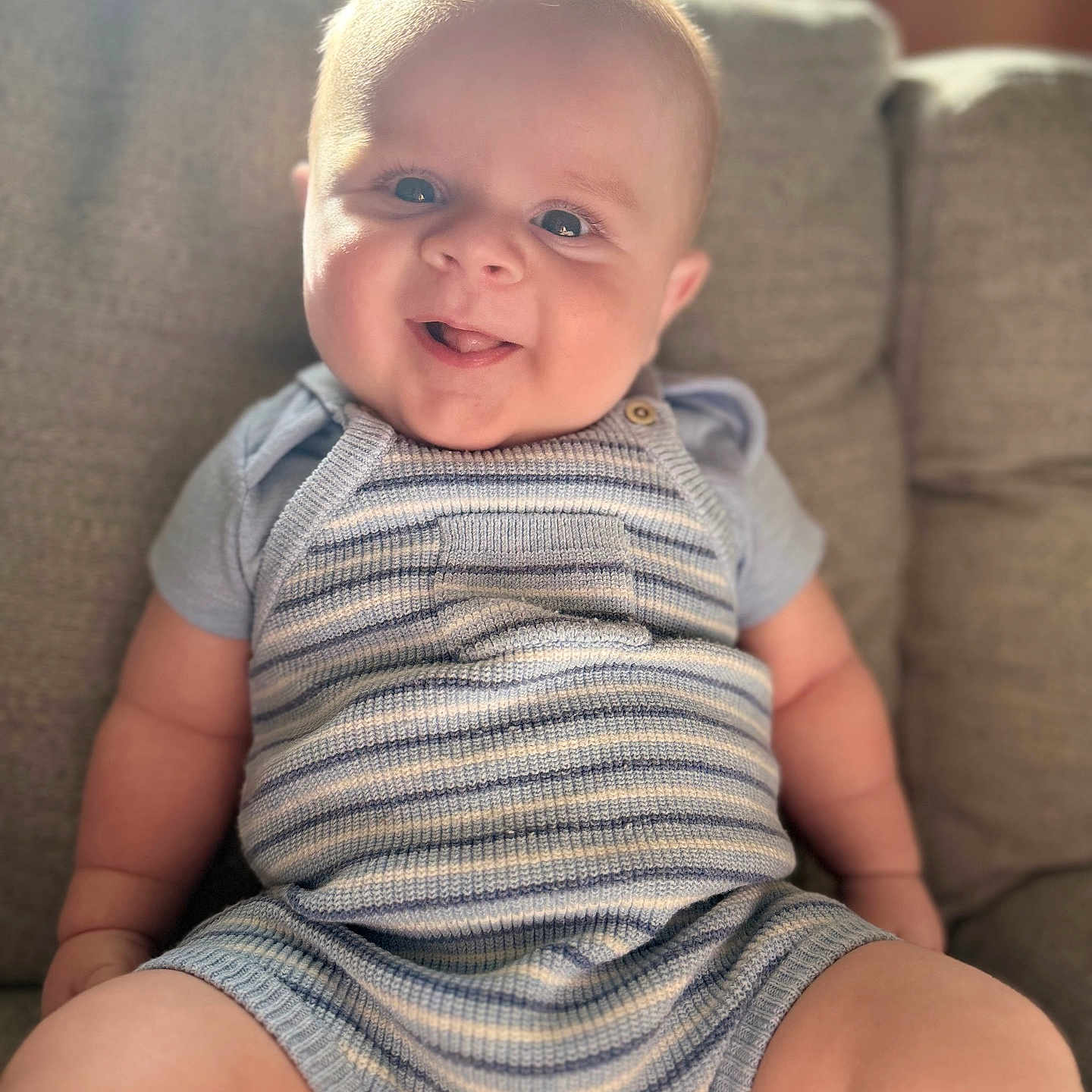 Alfie is registered to the contest to win money with this photo: baby, infant, smiling, cute, striped_clothing, romper, knitwear, seated, couch, indoor, soft_light, cheeks, tongue_out, happy, child, person, portrait, plush, cozy, comfortable