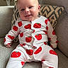 baby, child, smiling, onesie, red_tomato_pattern, couch, cushion, indoor, fan, happy, portrait, infant, clothing, fabric, head, face, person, seated, comfort, home