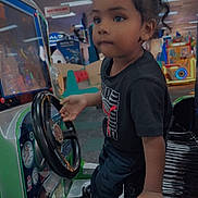 Edgar is registered to the contest to win money with this photo: toddler, child, arcade, gaming_machine, steering_wheel, indoor, playful, curly_hair, concentration, black_shirt, jeans, sneakers, hand, seat, car_game, amusement_center, bright_colors, entertainment, young_child, floor