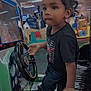 toddler, child, arcade, gaming_machine, steering_wheel, indoor, playful, curly_hair, concentration, black_shirt, jeans, sneakers, hand, seat, car_game, amusement_center, bright_colors, entertainment, young_child, floor
