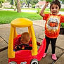 child, baby, toy_car, outdoor, play, grass, sidewalk, tree, leaf, fall_clothing, pink_shoes, red_car, yellow_roof, happy, smiling, person, nature, house, suburb, daylight
