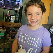 Kenslea is registered to the contest to win money with this photo: child, girl, smile, indoor, living_room, purple_shirt, stuffed_animal, unicorn, toy, furniture, television, cabinet, happy, person, brown_hair, casual_clothing, home, decor, couch, playful
