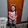 child, girl, smiling, pink_bow, backpack, minnie_mouse, shirt, shorts, black_shoes, carpet, door, laundry_basket, indoors, wooden_door, happy, hair, standing, person, home, clothing