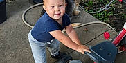 Jordan joined the competition — help win amazing prizes! toddler, child, tricycle, dog, outdoor, grass, potted_plants, concrete, sidewalk, toy, pet, smiling, casual_clothing, blue_shirt, gray_pants, black_shoes, sunlight, nature, playtime, friendship
