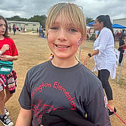 Charlee is registered to the contest to win money with this photo: child, girl, smiling, outdoor, school_event, face_paint, tshirt, blonde_hair, colorful_hair, people, cloudy_sky, grass, casual_clothing, person, portrait, happy, festival, community, daytime, event