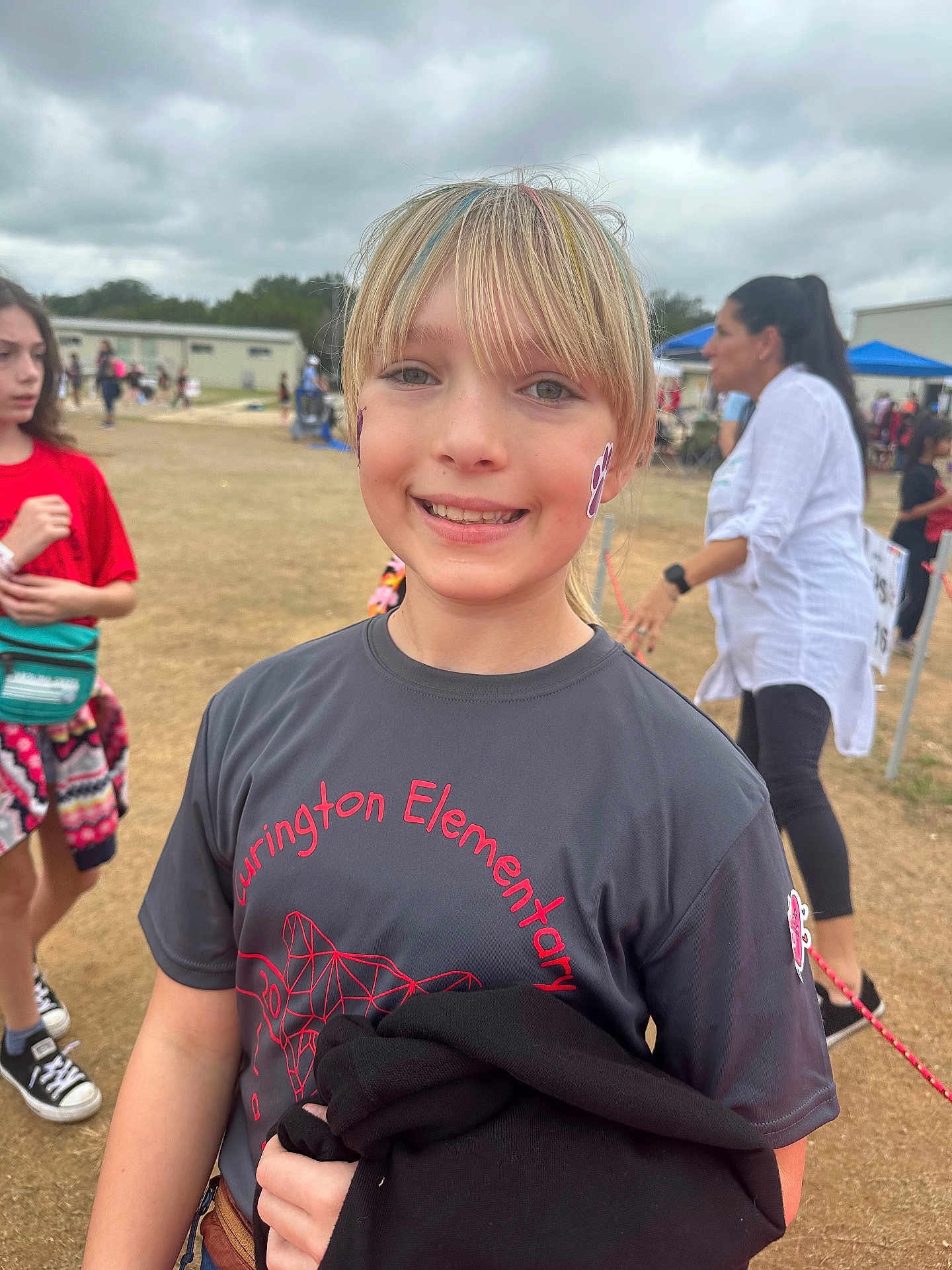 Charlee is registered to the contest to win money with this photo: child, girl, smiling, outdoor, school_event, face_paint, tshirt, blonde_hair, colorful_hair, people, cloudy_sky, grass, casual_clothing, person, portrait, happy, festival, community, daytime, event