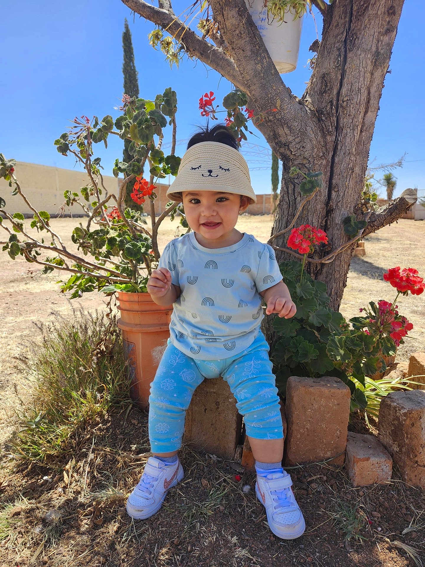 Hannah joined the competition — help win amazing prizes! child, toddler, hat, sun_visor, smiling, flowers, tree, outdoor, plants, blue_sky, grass, pot, sneakers, clothing, pants, shirt, nature, daylight, happy, person