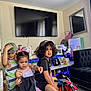 children, indoor, play, toy_motorcycle, cap, curly_hair, black_tshirt, striped_shirt, pigtails, living_room, television, couch, photos, table, curtains, floor, playful, kids, furniture, home